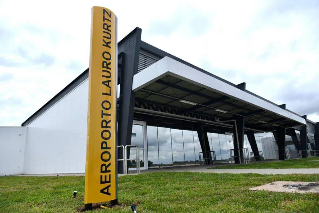 Uma placa amarela escrito 'Aeroporto Lauro Kurtz', na vertical. Levemente ao fundo, está o aeroporto.