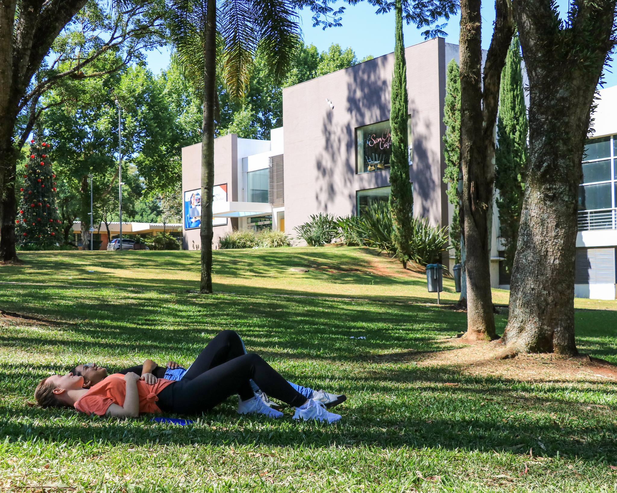 Duas pessoas deitadas em um gramado, na frente do centro de convivências da UPF, um prédio cinza e branco. Há algumas arvores.